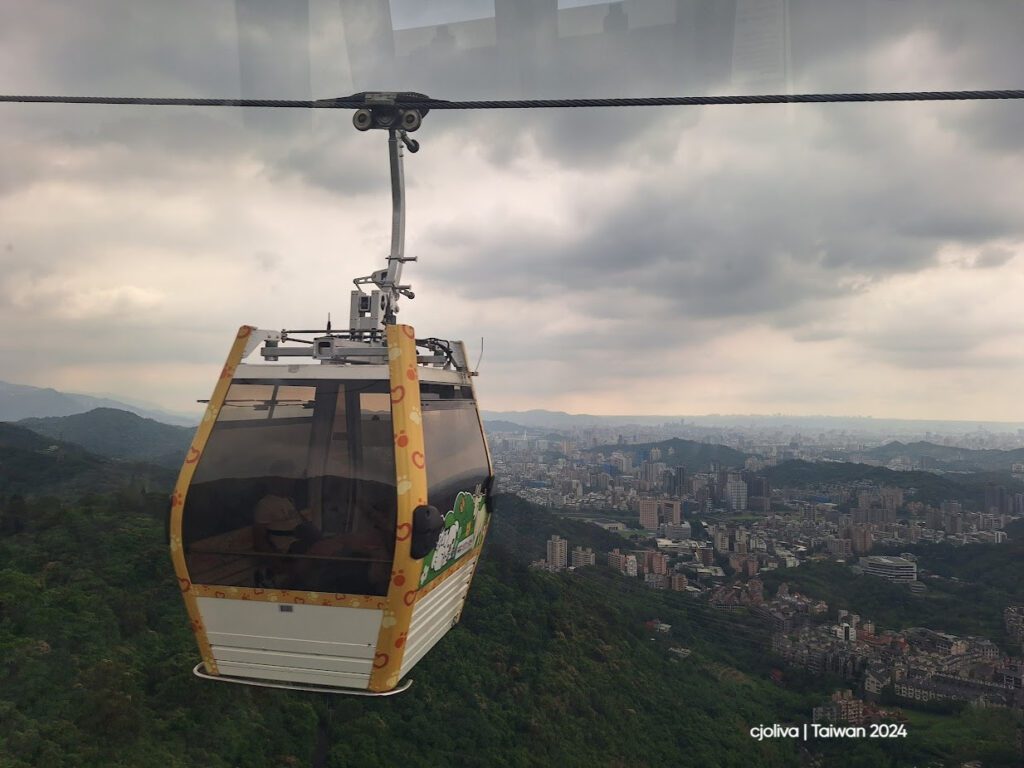 The Maokong Gondola cable car is suspended above green mountains with the Taipei cityscape in the background under a cloudy sky.