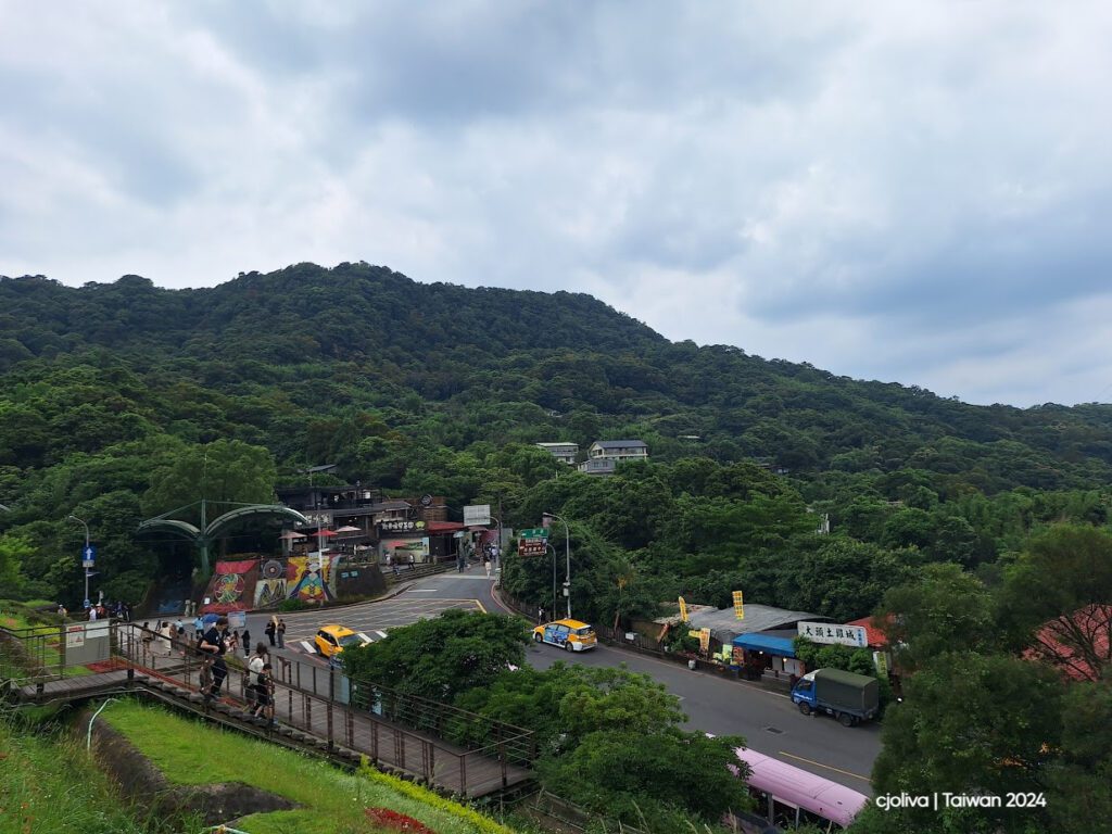 Hillside town at Maokong in Taipei with murals, a road lined with taxis, pedestrians on the walkway, local businesses, and a dense green forest under a cloudy sky.