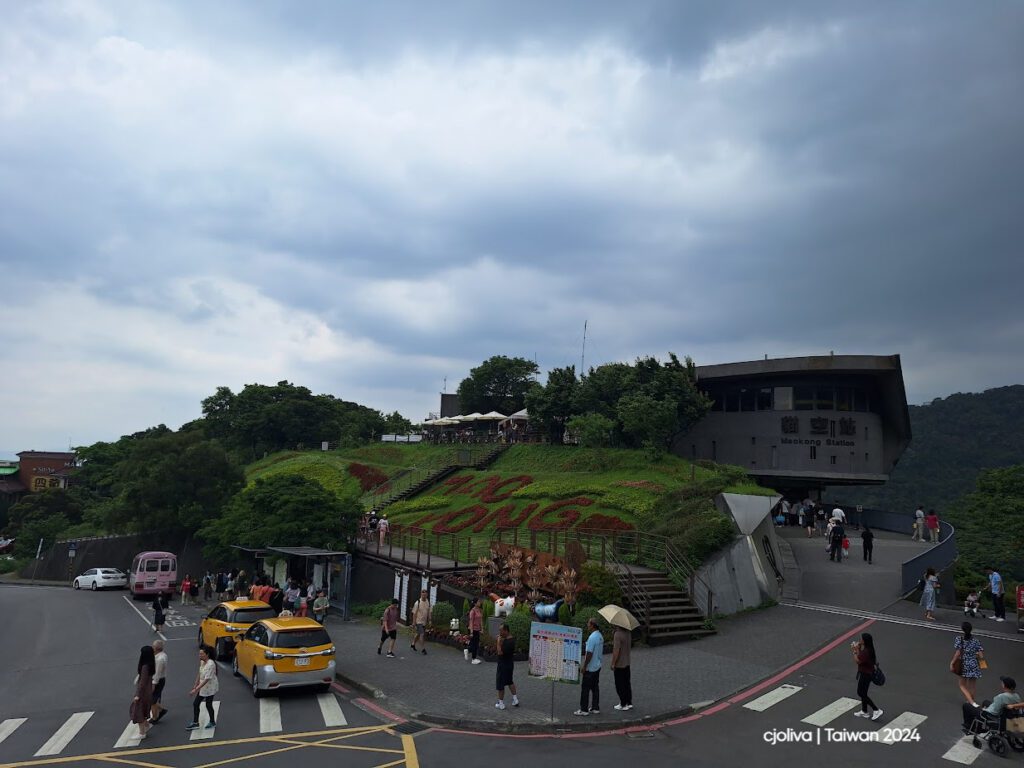 Maokong Station in Taipei on a hillside with landscaped greenery spelling ‘Maokong,’ people walking, taxis and buses on the road, under a cloudy sky.