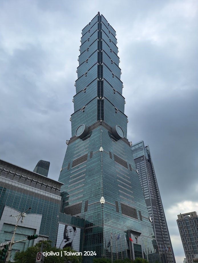 The Taipei 101 skyscraper in Taiwan, with a bamboo-inspired design, a green glass facade, tiered sections, and flags at the base under a cloudy sky.