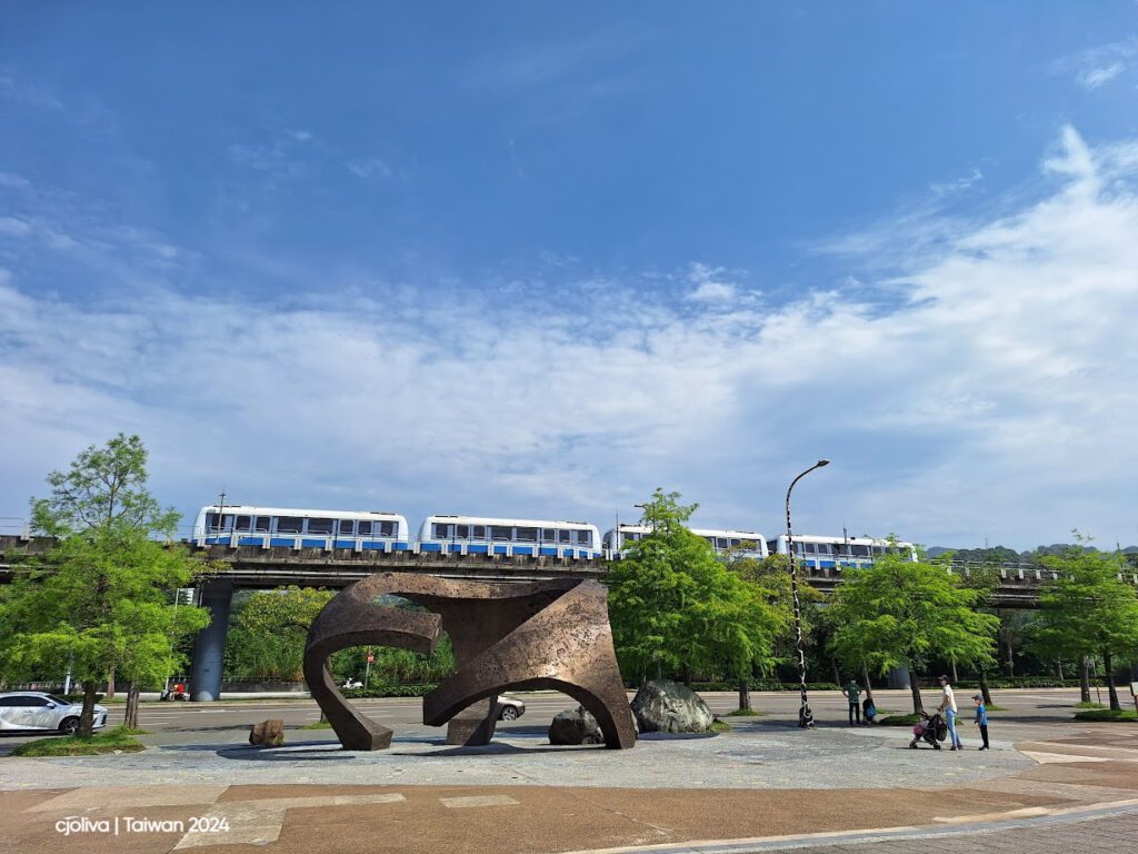 A Brown Line train passing behind an abstract bird sculpture in Taipei, with trees, a clear sky, and people in a public space near Taipei Zoo.
