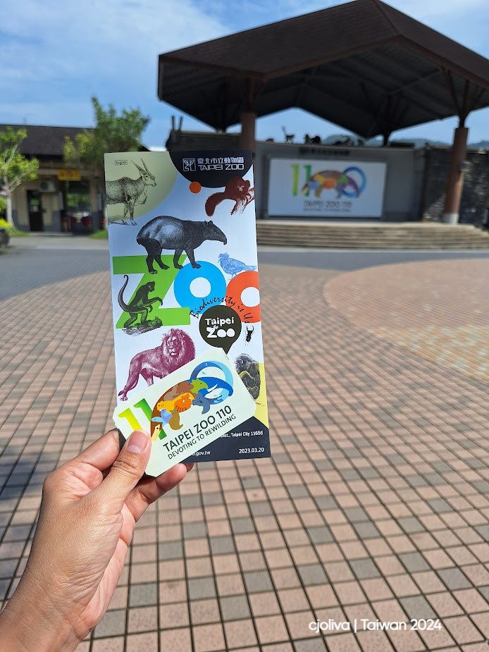 Colorful Taipei Zoo brochure is held in front of the zoo entrance, featuring animals like tapirs, lions, and monkeys with biodiversity and conservation themes.