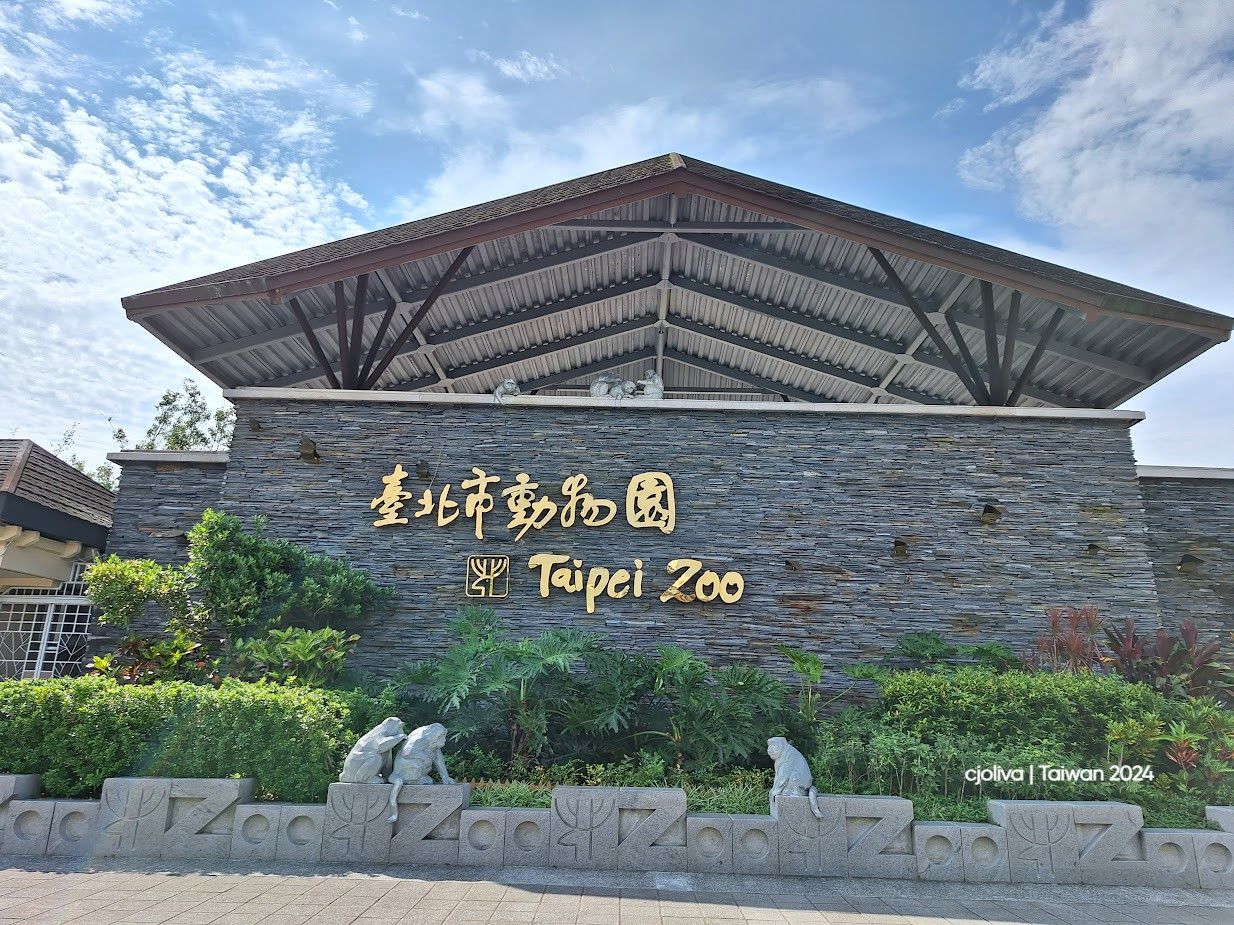 Entrance to Taipei Zoo in Taiwan with a stone wall, golden Chinese and English text, animal statues, and a slanted metal roof surrounded by greenery.