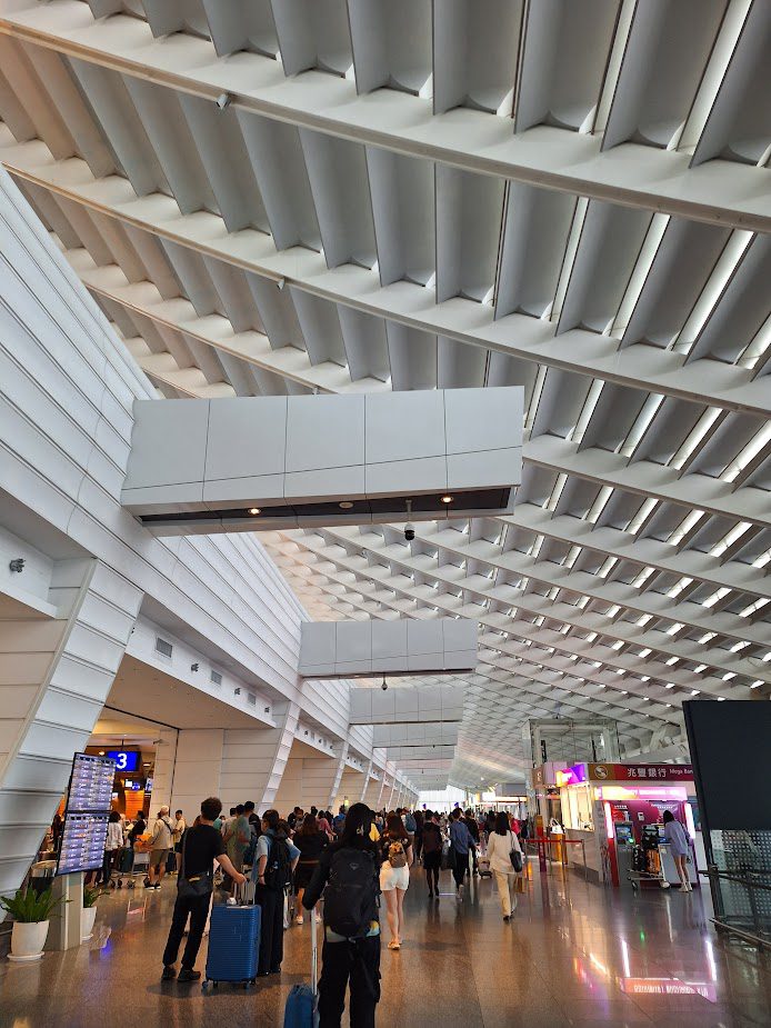 Modern airport terminal in Taiwan with geometric ceiling panels, skylights, travelers with suitcases, flight information display, and China Airlines counters.