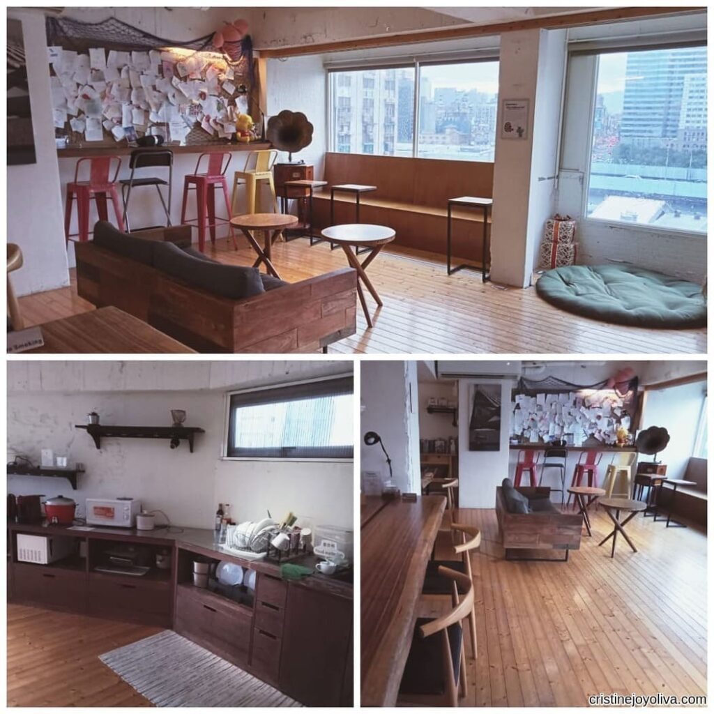 A cozy hostel lounge in Taipei with a wooden bench, corkboard wall, and city skyline view through large windows.