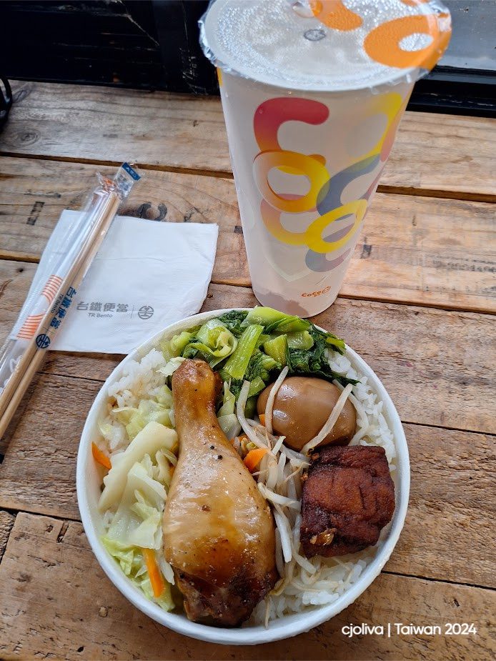 A Taiwan Railway bento meal with rice, chicken drumstick, braised egg, pork belly, vegetables, and a CoCo drink on a rustic wooden table.