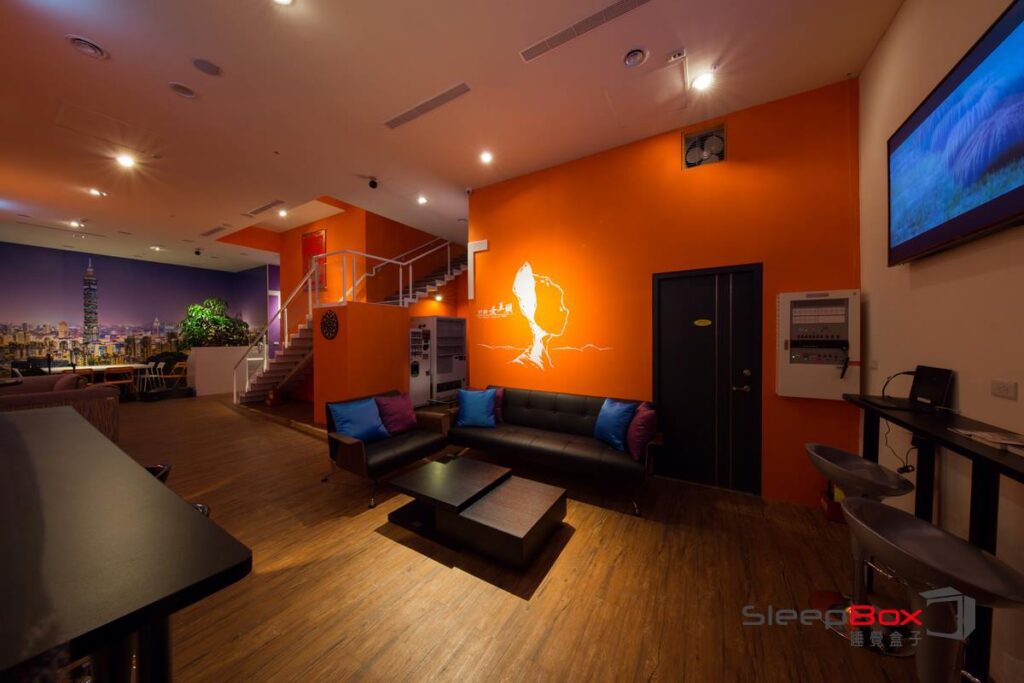 SleepBox lounge in Taipei with orange and white walls, a city skyline mural, black leather sofas with colorful cushions, a staircase, a vending machine, a dartboard, and warm lighting.