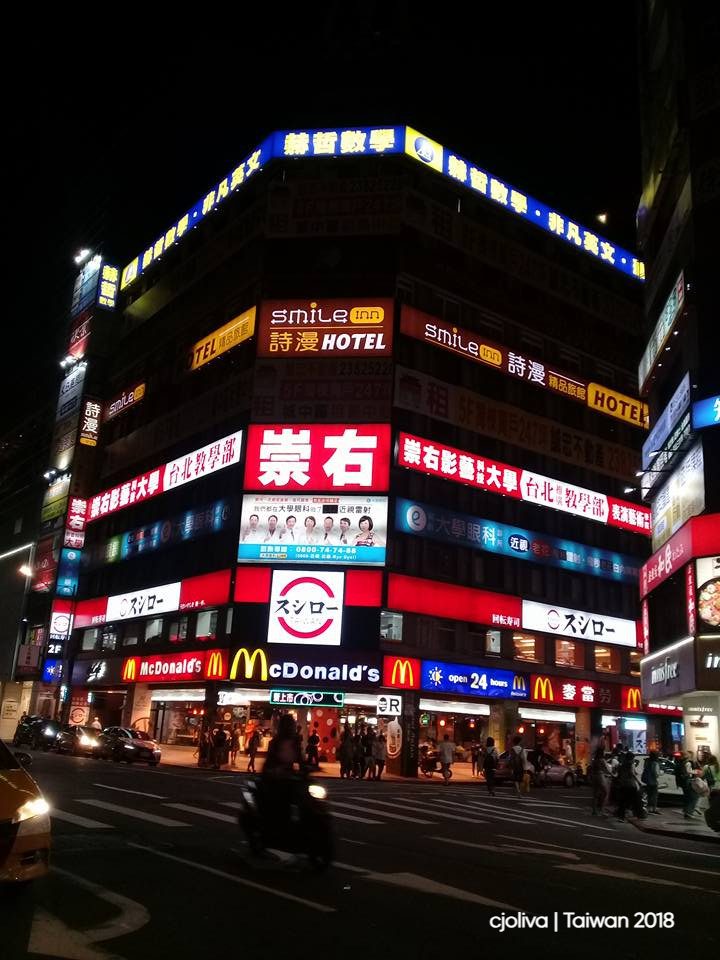 Taipei street corner at night with a multi‑story building covered in neon signs, McDonald’s open 24 hours, Smile Inn hotel, Sushiro sushi chain, and pedestrians with scooters.