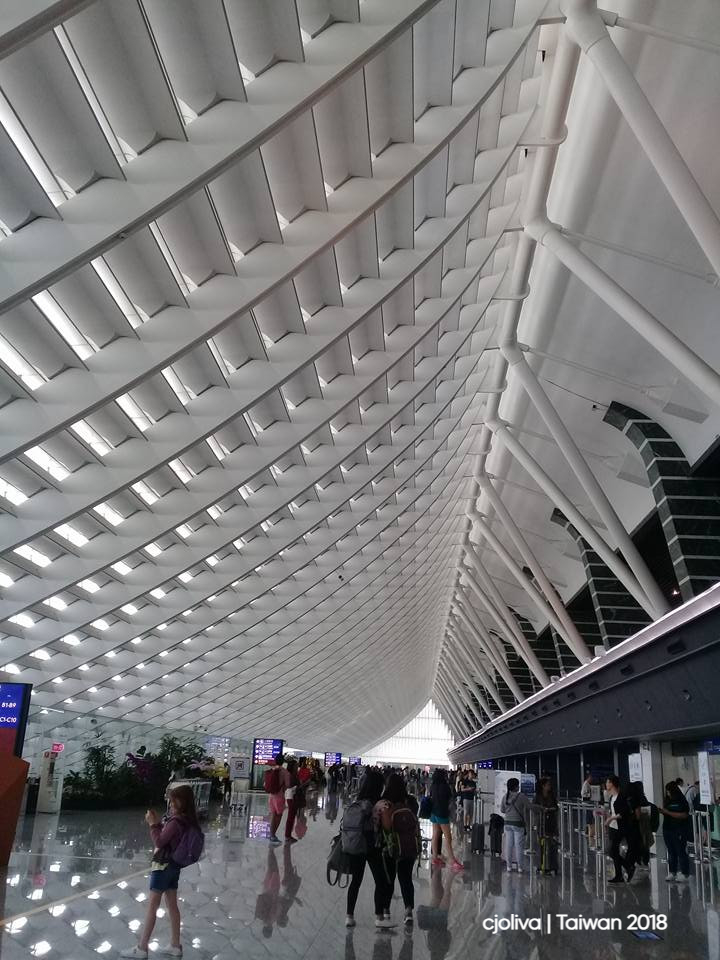 Modern airport terminal in Taiwan with wave‑like ceiling panels, reflective floor, travelers with luggage, and digital flight information displays.