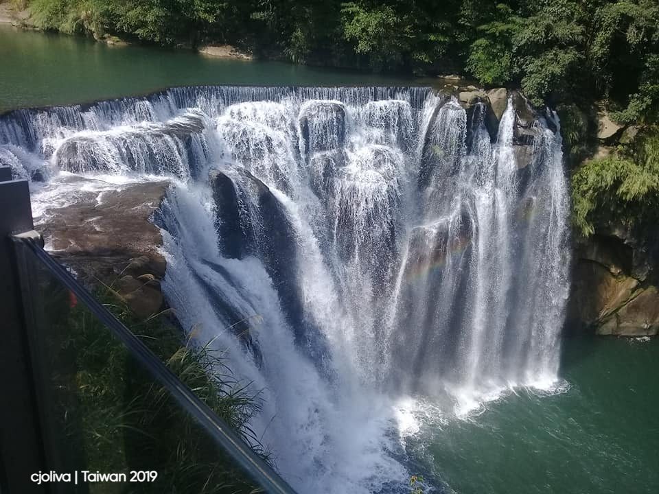 The multi‑tiered Shifen Waterfall in Taiwan cascades into a greenish‑blue pool, surrounded by lush greenery, with a faint rainbow forming in the mist near the base.