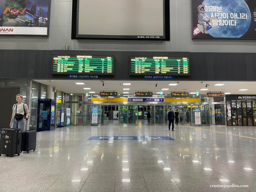 Interior view of Busan Station concourse in South Korea at 6:16 AM. Digital departure screens show KTX and SRT trains to Seoul and Suseo. A traveler with a suitcase is in the foreground of the bright, modern train terminal.