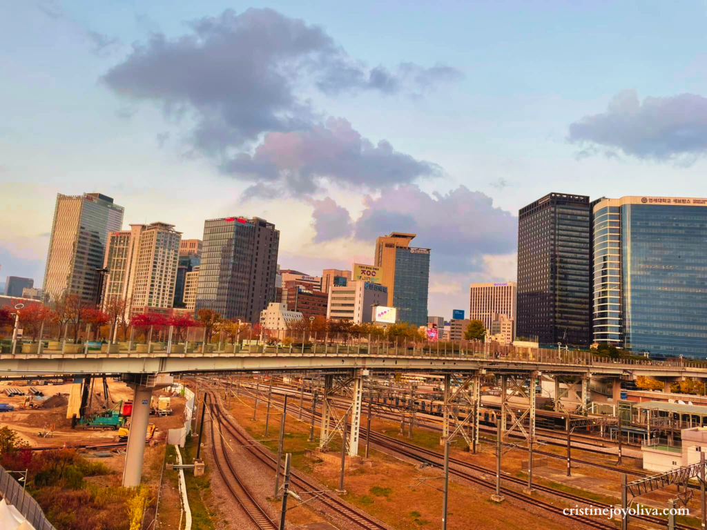 View from Seoullo 7017 sky garden looking toward the Seoul city skyline. An elevated pedestrian walkway crosses over multiple train tracks with modern glass skyscrapers and a colorful sunset sky in the background.