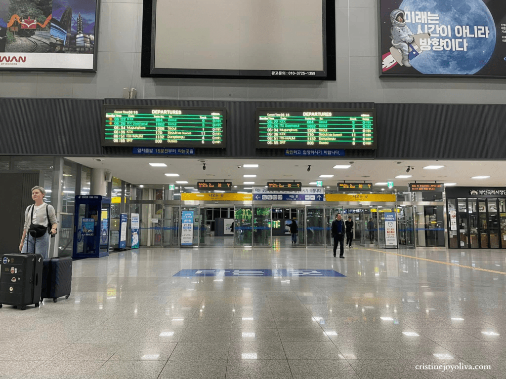 Interior of Busan Station showing digital departure boards, travelers with luggage, and entrance doors.