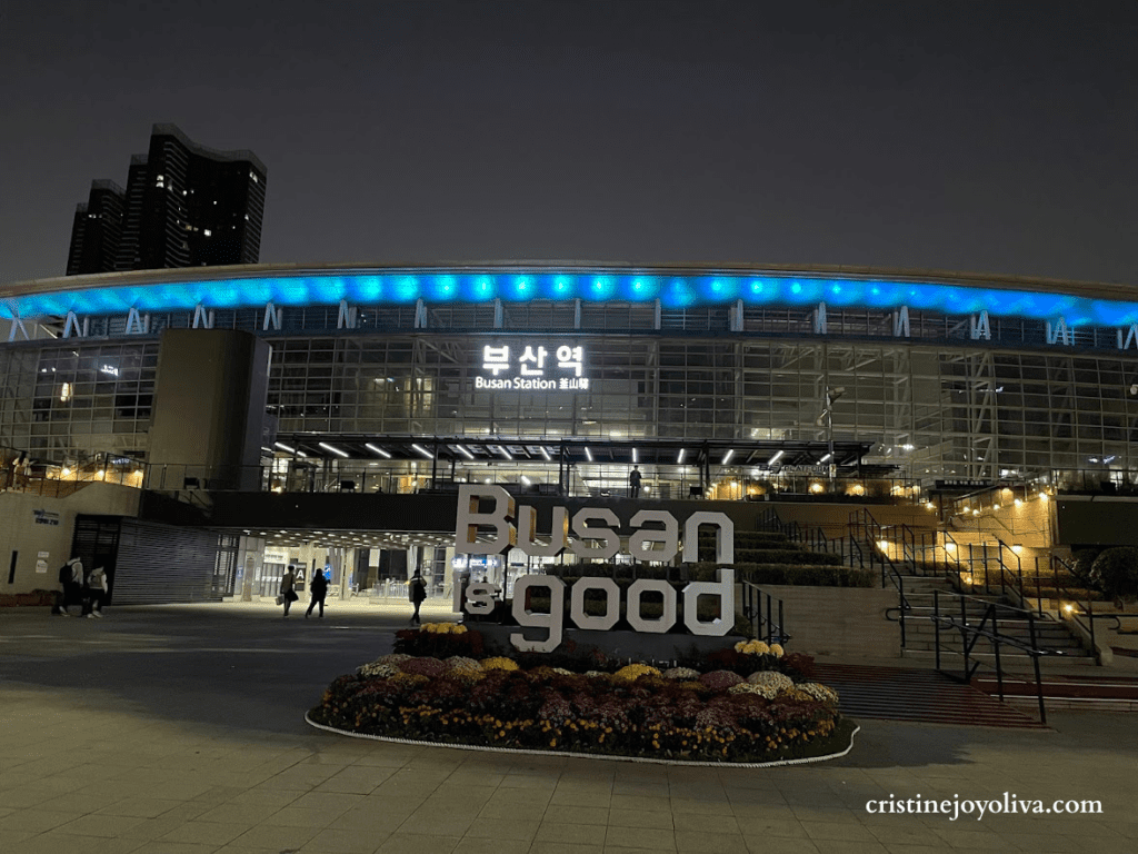 Night view of Busan Station in South Korea with illuminated "Busan is good" sign and blue lights on the station roof.