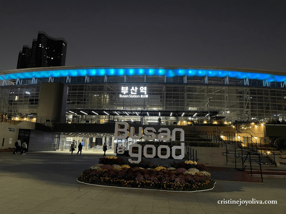 Night view of Busan Station in South Korea with illuminated "Busan is good" sign and blue lights on the station roof.
