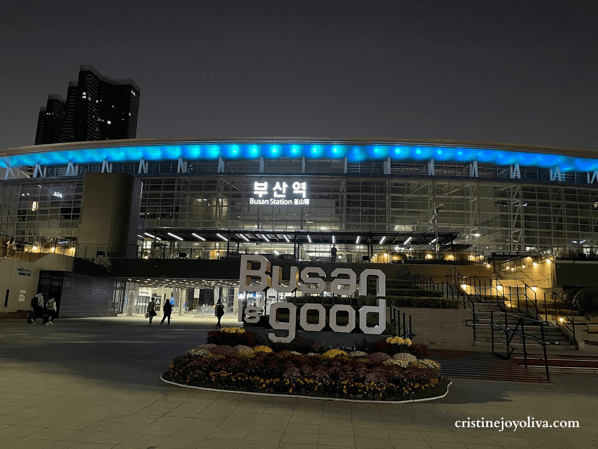Night view of Busan Station in South Korea with illuminated "Busan is good" sign and blue lights on the station roof.