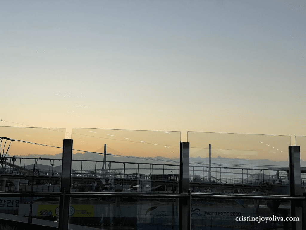 The sunrise sky is seen through glass panels at Busan Station with a distant bridge and mountains in the background.