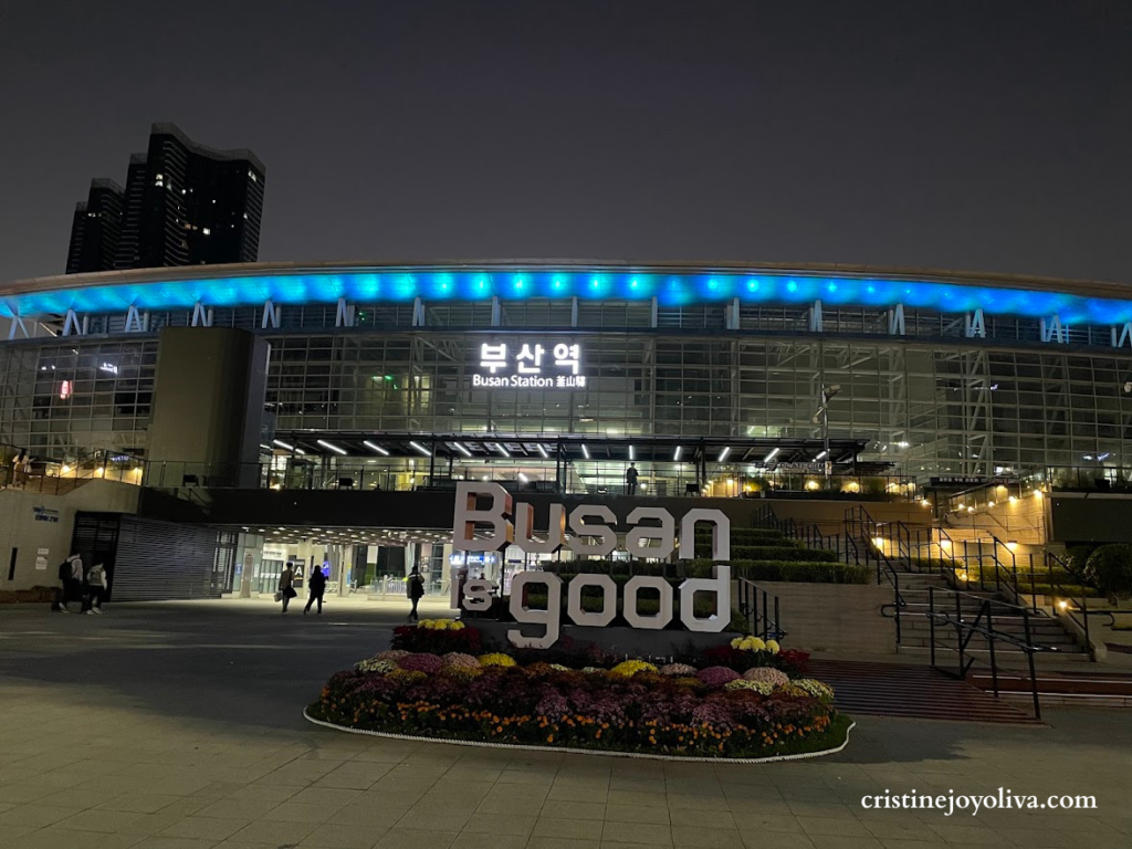 Night view of the modern Busan Station exterior in Busan, South Korea, featuring blue architectural LED lights. In the foreground, the large white text sculpture reads 'Busan is good' above a bed of colorful flowers. Pavement and people are visible in the plaza.