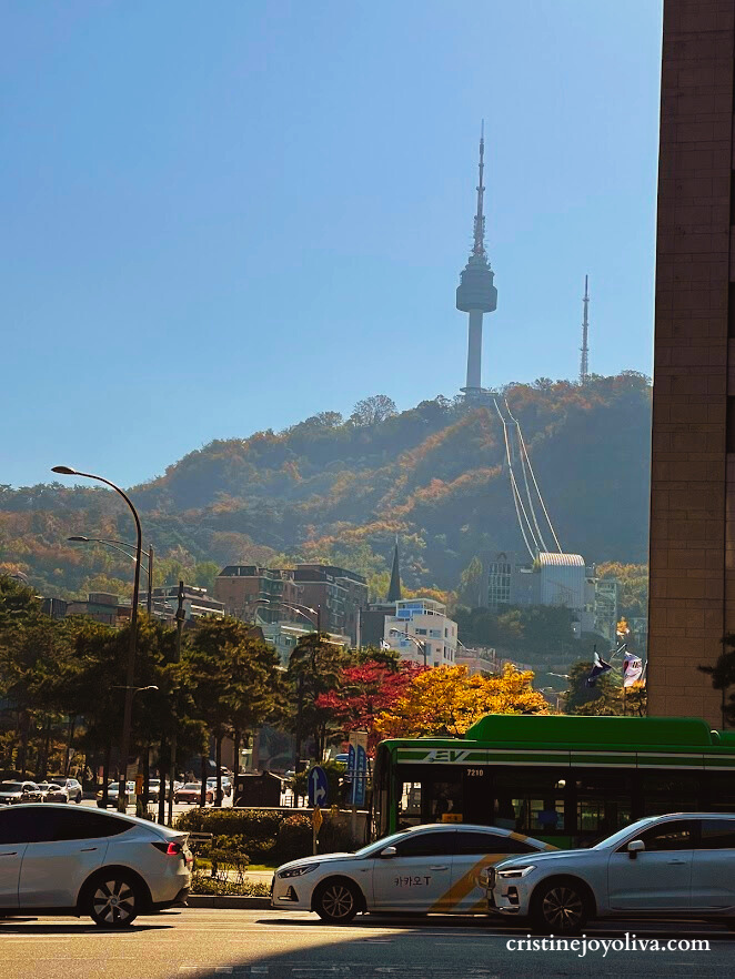 A low-angle view of N Seoul Tower on Namsan Mountain under a clear blue sky. The photo shows the Namsan Cable Car tracks, autumn trees on the hill, and a busy street with a green city bus and white taxis in the foreground.