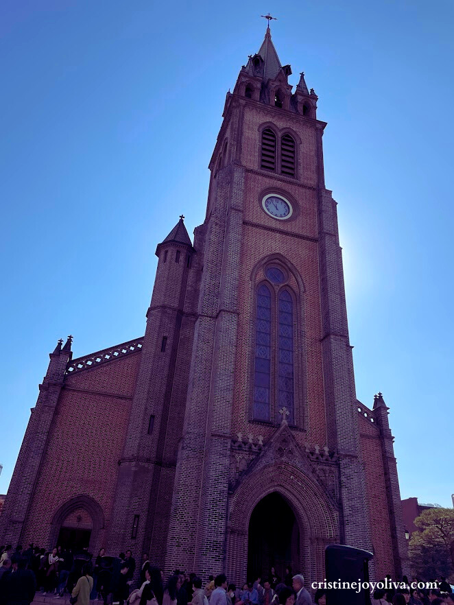 A low-angle view of the historic Myeongdong Cathedral in Seoul, South Korea. The red brick Gothic Revival church features a tall clock tower and central spire against a bright blue sky with people gathered at the entrance.