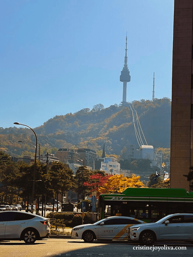 View of Namsan Seoul Tower on a hill with colorful autumn trees and busy city traffic on the street below.