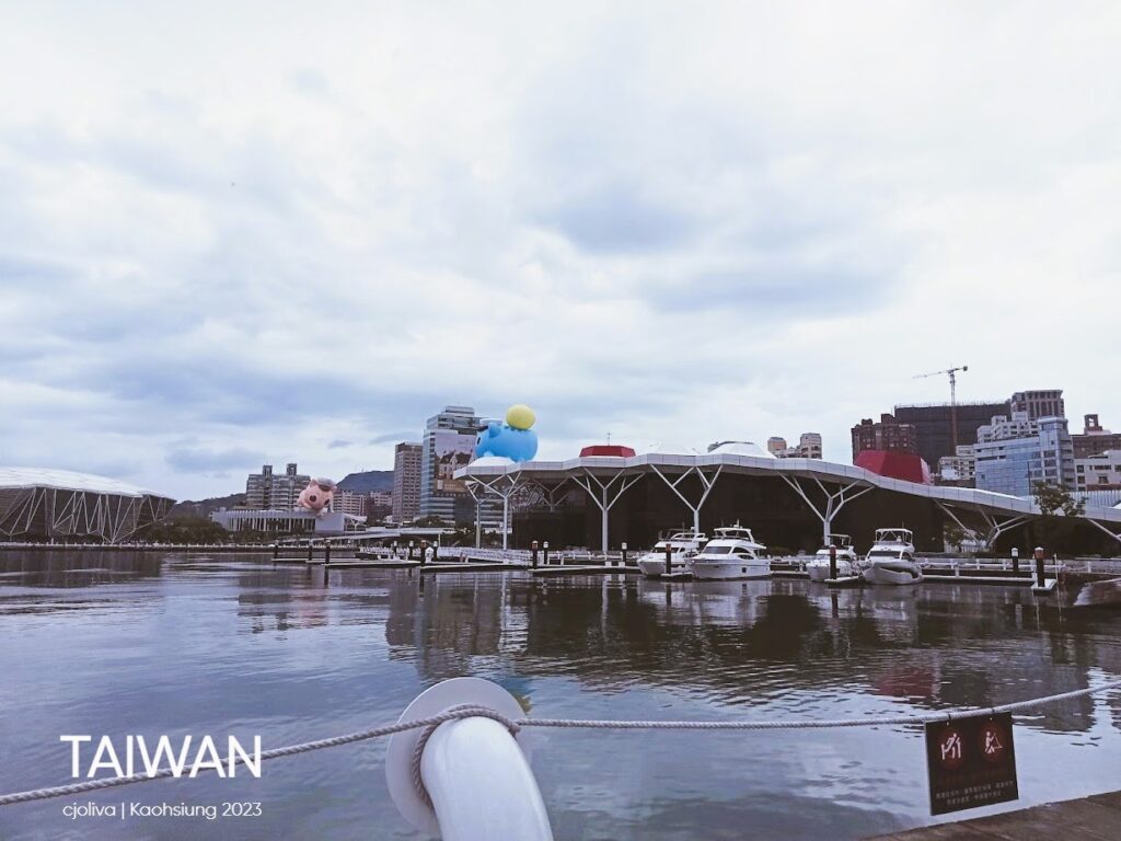 Scenic view of Kaohsiung, Taiwan, featuring a harbor with large character art installations (like a giant blue sculpture) and a modern exhibition center on a cloudy day.