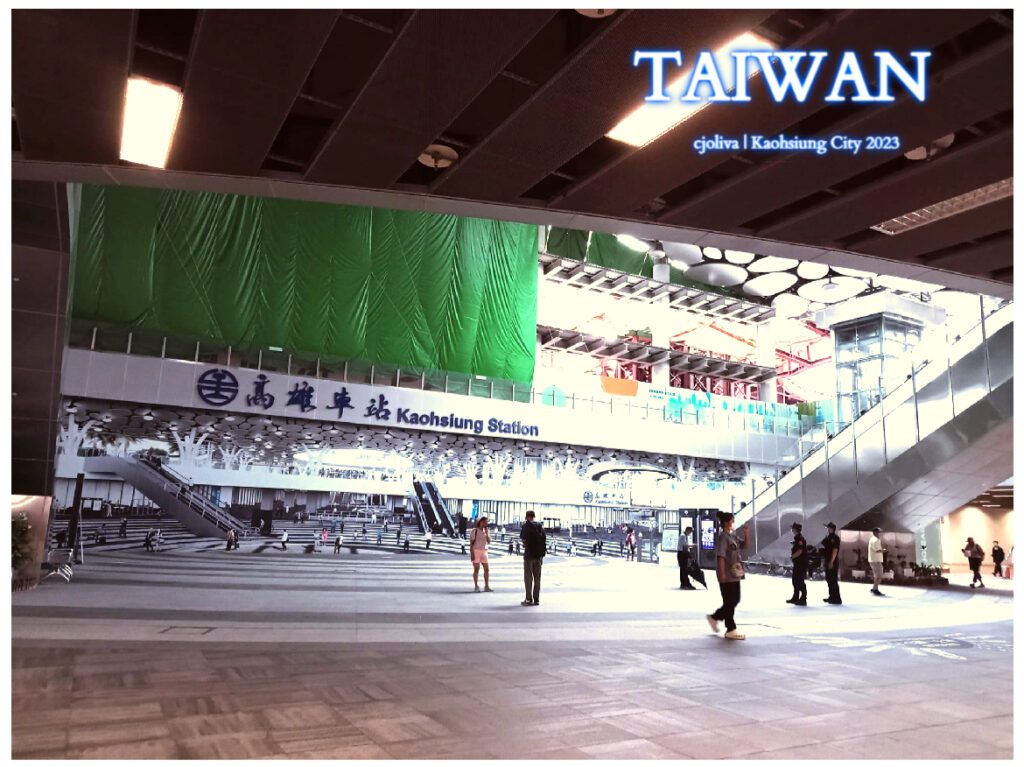 A wide-angle view of the modern Kaohsiung Station interior in Taiwan, featuring a large mural of the station's concourse, sleek architectural lines, and commuters walking through the bright, high-ceilinged transit hub.