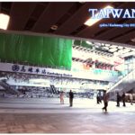 A wide-angle view of the modern Kaohsiung Station interior in Taiwan, featuring a large mural of the station's concourse, sleek architectural lines, and commuters walking through the bright, high-ceilinged transit hub.