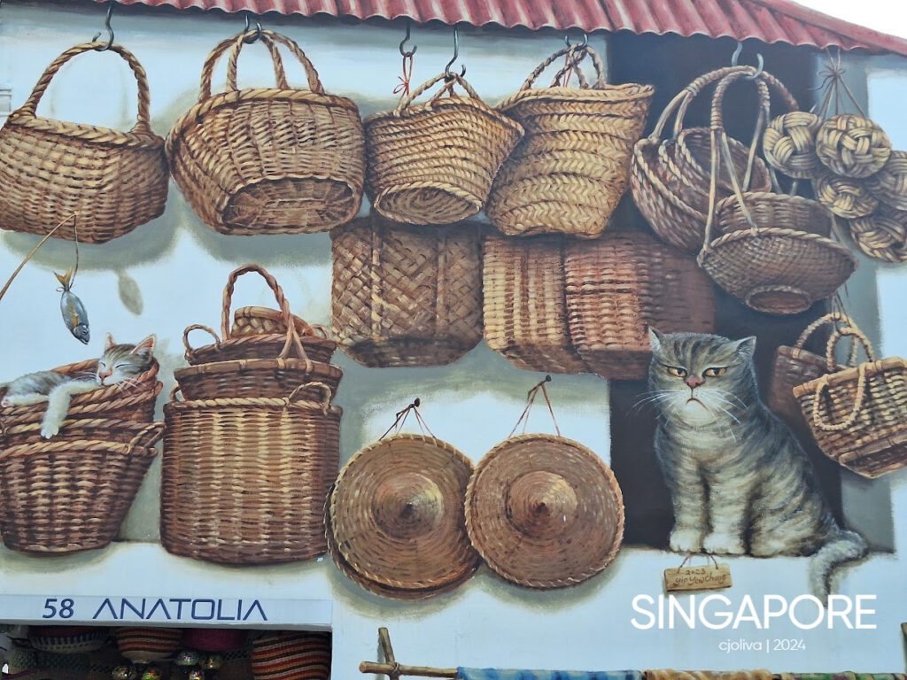 Mural in Singapore depicting woven baskets, straw hats, and two painted cats—one sleeping in a basket and one sitting upright—beneath a red-tiled roof.