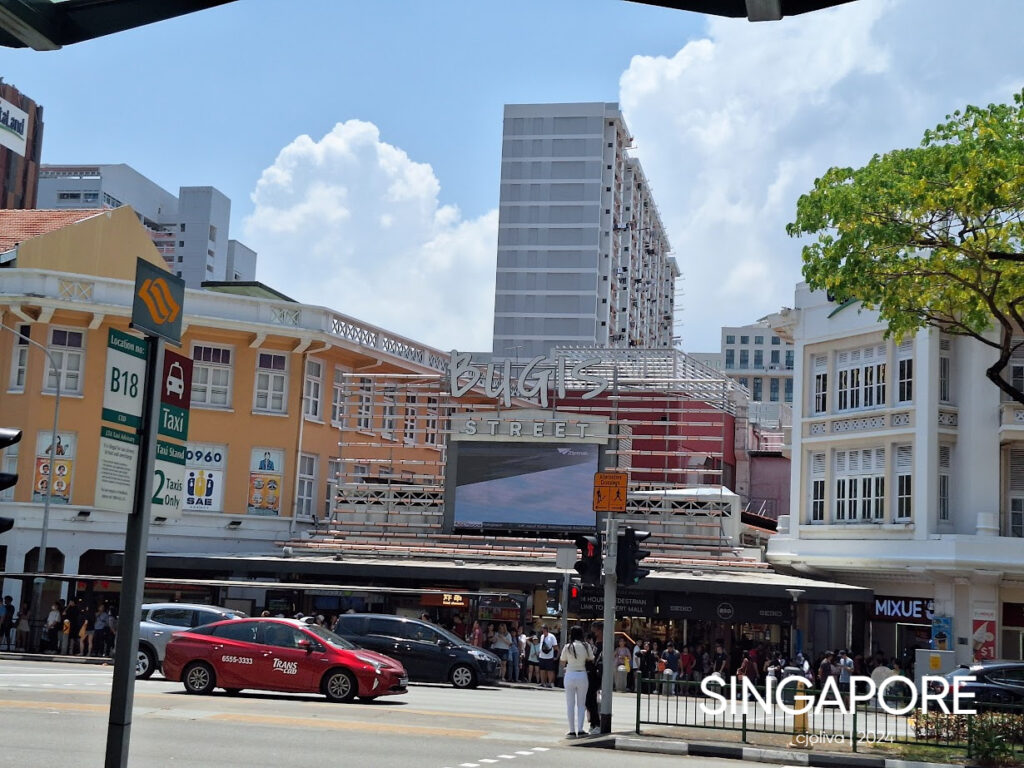 Busy Bugis Street entrance in Singapore with colonial-style buildings, a large sign, a red taxi, pedestrians, and a modern high-rise in the background.