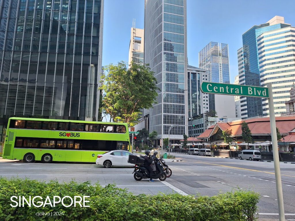Busy Singapore street at Central Boulevard with a green double-decker bus, modern skyscrapers, and a traditional red-roof building.