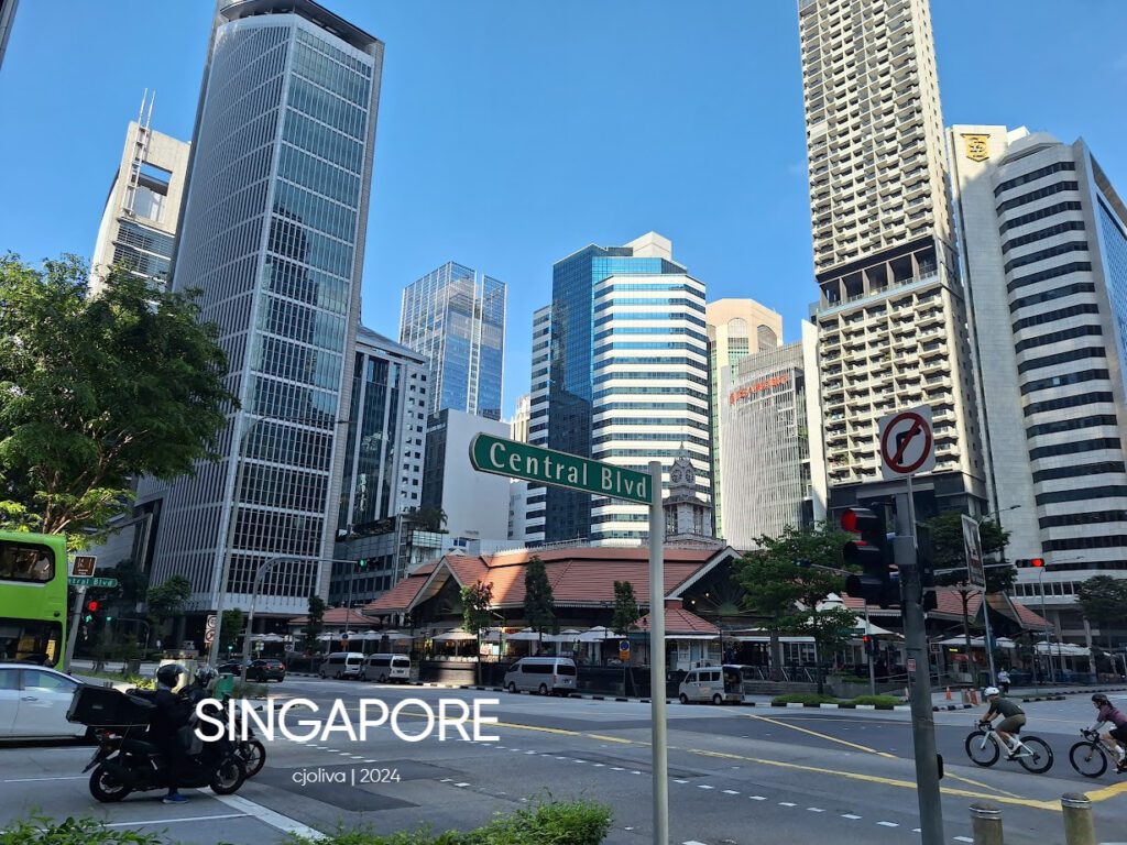 Busy intersection at Central Boulevard in Singapore with modern skyscrapers, traditional red-roof buildings, cars, motorcycles, bicycles, and a green double-decker bus.