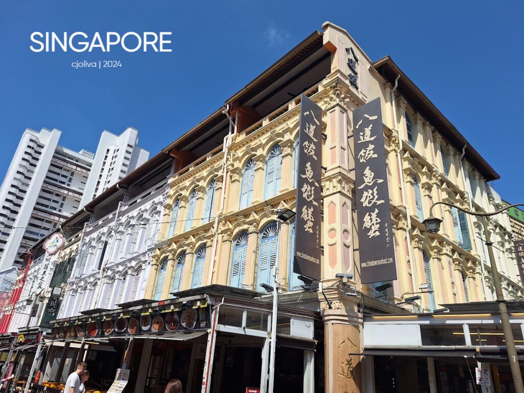 Colorful colonial-style shophouses in Singapore’s Chinatown with pastel facades, arched windows, Chinese signage, and outdoor seating at a seafood restaurant under a clear blue sky.