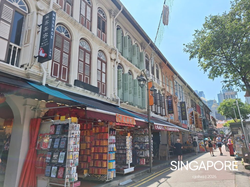 Vibrant Singapore shophouses with pastel facades, arched windows, souvenir shops, and pedestrians along a lively Chinatown street with modern high-rises in the background.