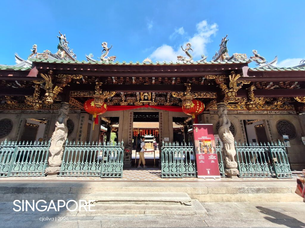 Ornate Chinese temple entrance in Singapore with dragon roof sculptures, golden carvings, red lanterns, and green wrought iron gate under a clear blue sky.