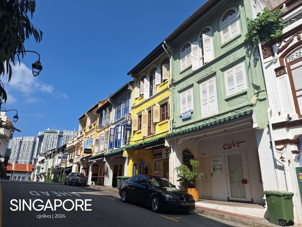 Row of colorful shophouses in Singapore with shuttered windows and decorative moldings, alongside modern skyscrapers in the background.