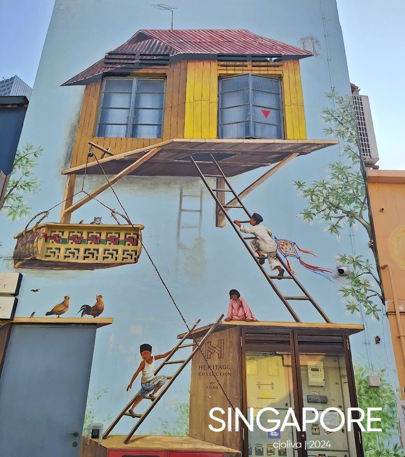 Large mural in Singapore depicting a traditional wooden stilt house with children, chickens, and bamboo plants, blending cultural heritage with urban art.