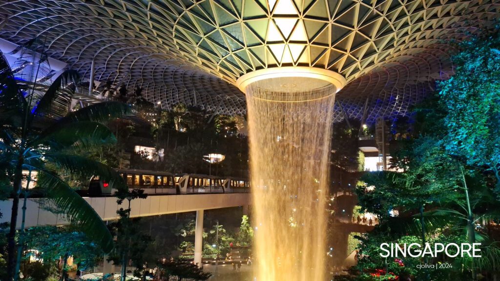 Interior of Jewel Changi Airport in Singapore featuring the Rain Vortex waterfall descending from a glass dome, surrounded by lush greenery and a passing train.