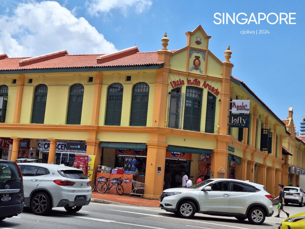 Colonial-style Little India Arcade in Singapore with yellow and orange façade, green shutters, red-tiled roof, and street life with cars, bicycles, and pedestrians.