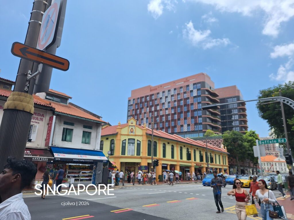 Colorful two-story shophouses in Singapore with shuttered windows, tiled roofs, Maxi-Cash pawn shop signage, and pedestrians along a lively street under a partly cloudy sky.
