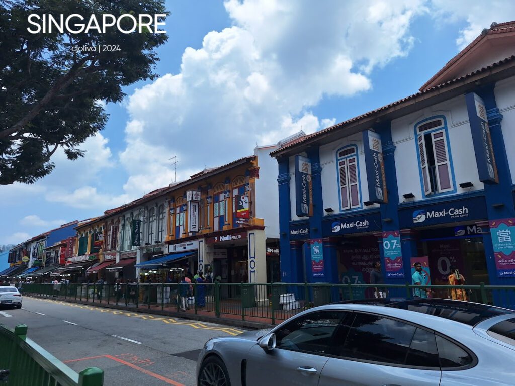 Colorful two-story shophouses in Singapore with shuttered windows, tiled roofs, Maxi-Cash pawn shop signage, and pedestrians along a lively street under a partly cloudy sky.