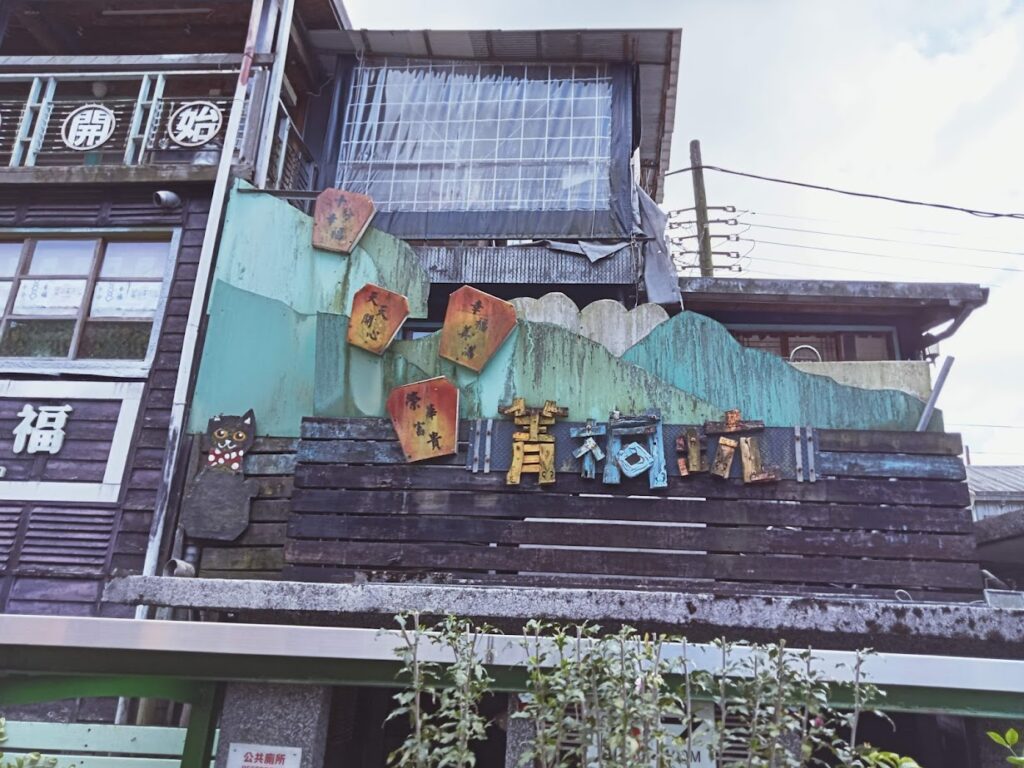 Rustic building in Jingtong, New Taipei, Taiwan, with weathered wood and metal textures, colorful wooden panels, hexagonal plaques with Chinese characters, a cat‑shaped figure, circular signs, and decorative signage reading “Jingtong Little Town.”