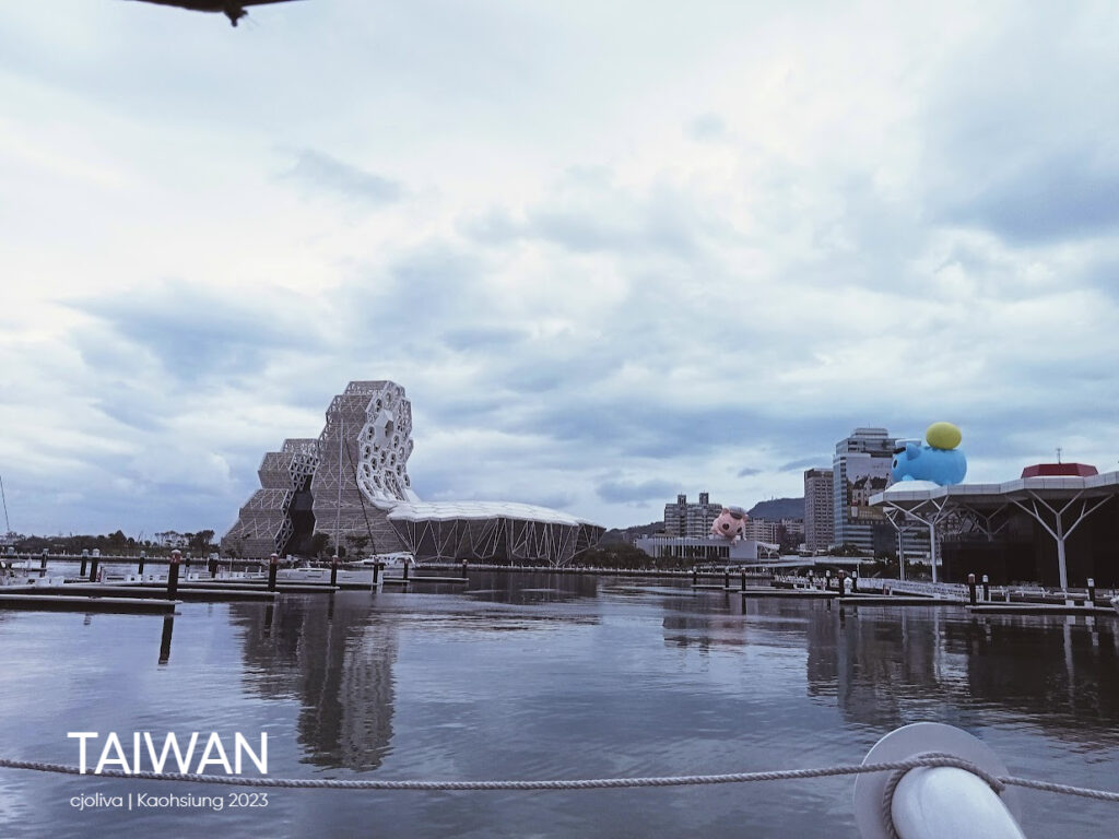 Kaohsiung Music Center in Taiwan with a honeycomb‑like geometric design beside a calm waterfront, reflections of architecture and sky, city buildings in the background, and colorful inflatable figures under an overcast sky.