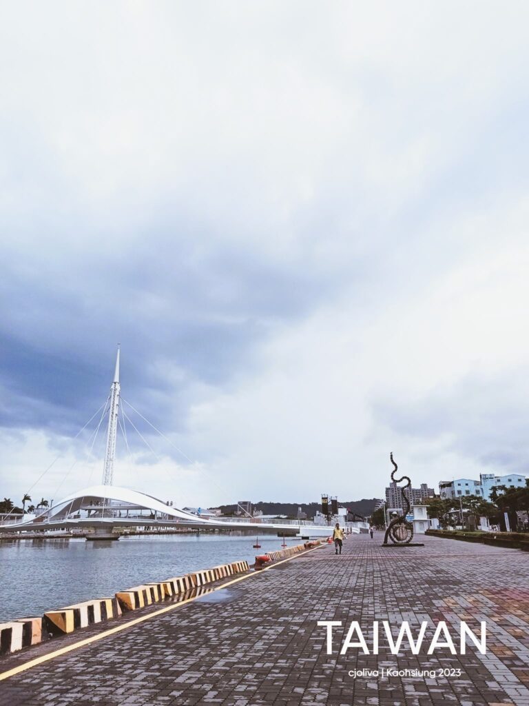Waterfront in Kaohsiung, Taiwan featuring modern white cable‑stayed pedestrian bridge with tall mast and curved deck, paved walkway with striped barriers and buoys, person walking toward abstract twisting metal sculpture, mid‑rise buildings and greenery under cloudy sky.