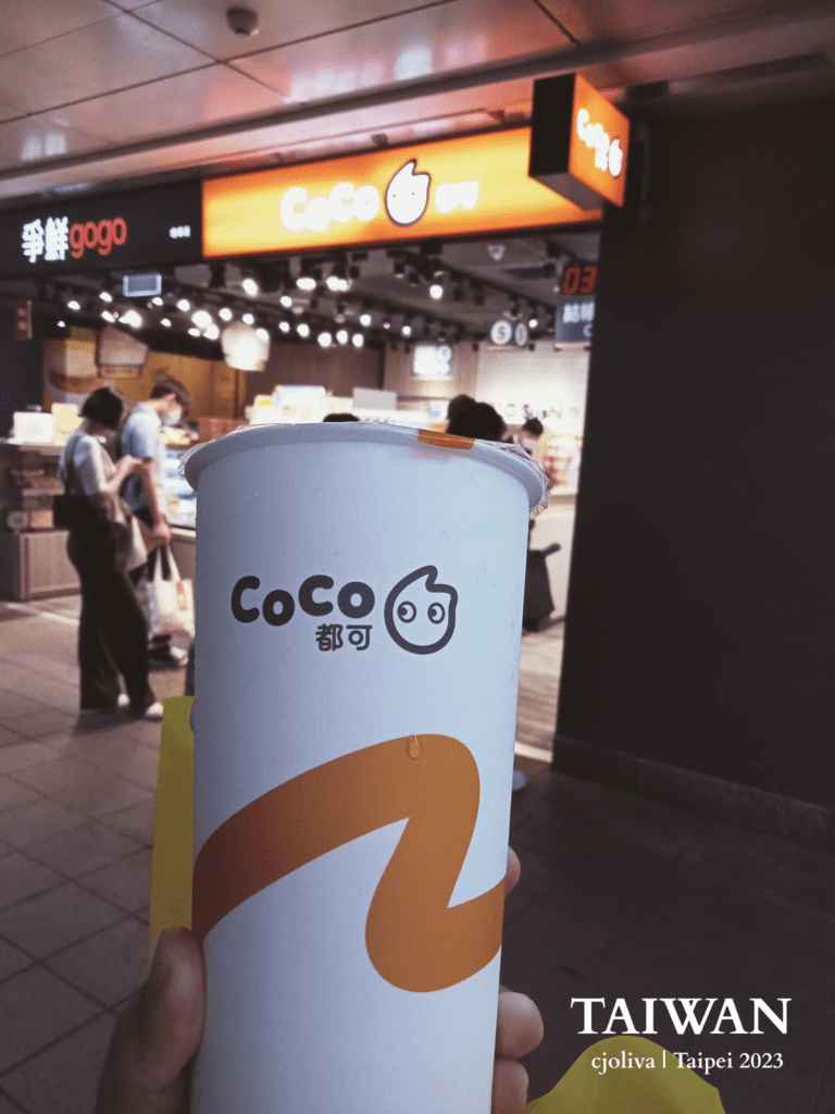 A hand holding a signature white and orange CoCo Fresh Tea & Juice cup in a Taipei metro station, with a blurred CoCo storefront and commuters in the background.