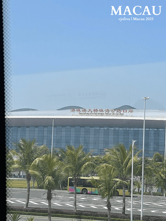 A view through a window of the Hong Kong-Zhuhai-Macau Bridge Zhuhai Port building under a clear blue sky, with palm trees and a yellow bus in the foreground.