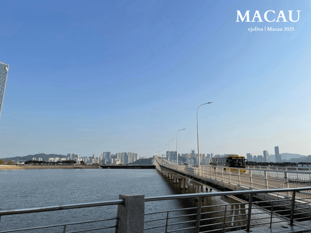 A yellow public bus drives across the Governor Nobre de Carvalho Bridge in Macau, with a wide view of the city skyline and blue water under a clear sky.