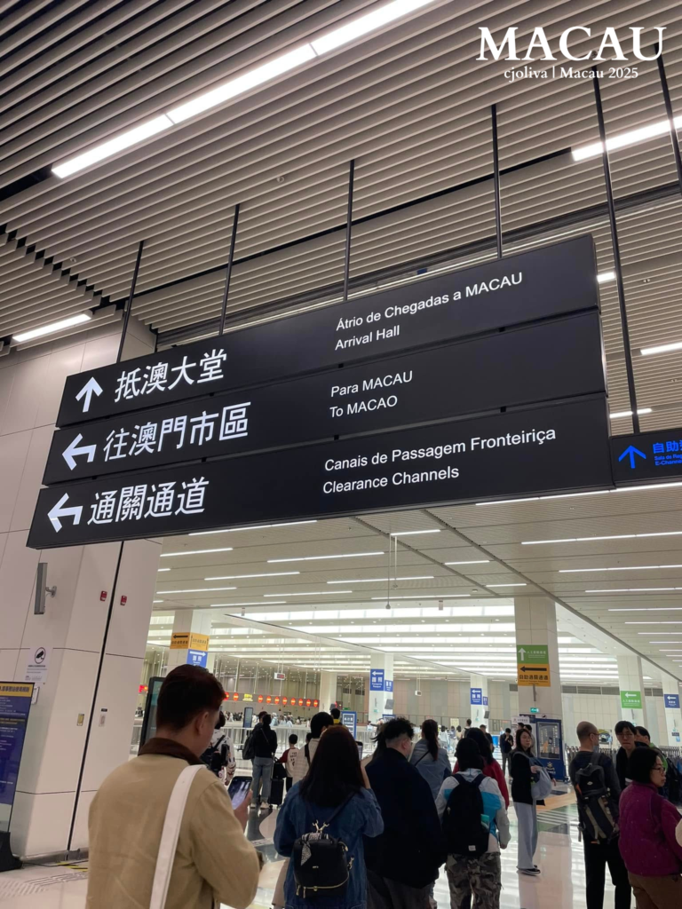 A view inside the modern Macau Boundary Crossing Facilities arrival hall. Black directional signs in Chinese, Portuguese, and English point toward the Arrival Hall, Macau city center, and Clearance Channels, with travelers walking below.
