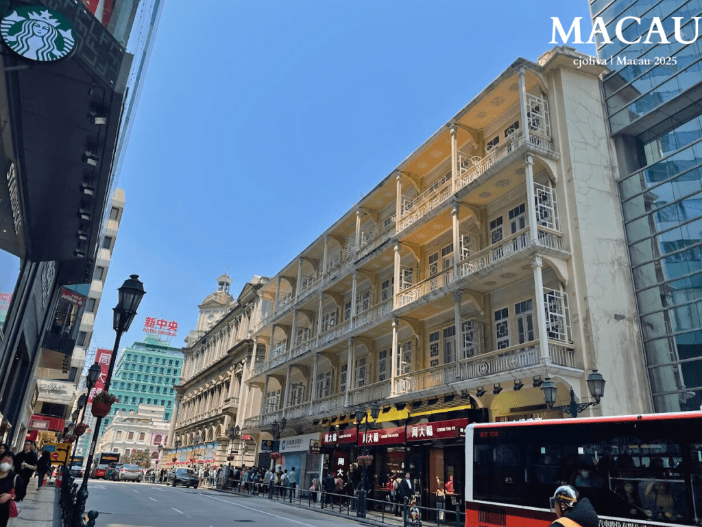 A street view of Avenida de Almeida Ribeiro (San Ma Lo) in Macau, featuring a grand four-story yellow colonial-style building with intricate white balconies. A red public bus and pedestrians are visible on the sunlit street.