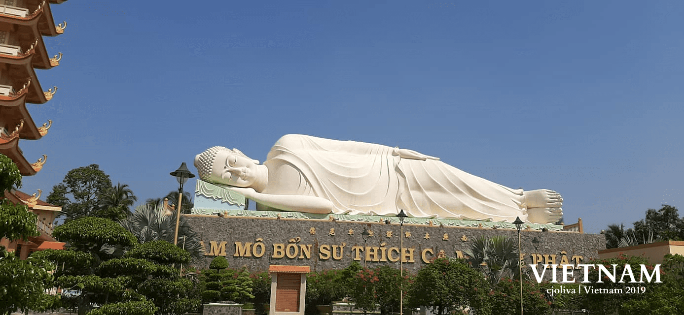 Reclining Buddha statue at a temple visited on the Mekong River Delta Tour, surrounded by lush greenery and clear blue sky.